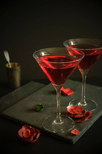 Two cosmopolitan cocktails in a triangular glass. In the background an old silver bowl and scattered rose petals. Dark luxury background.High resolution. - Foto, afbeelding