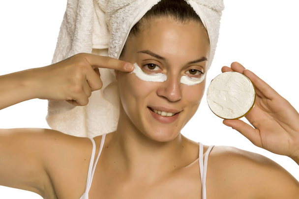 Joven mujer sonriente aplicando crema facial bajo sus ojos sobre fondo blanco
  - Foto, Imagen