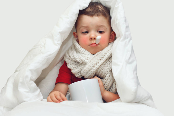 A little cute boy gets sick and measures the temperature with an oral thermometer and sits covered with a blanket and with a large knitted scarf around his neck.He squinted and looks at the thermomete - Photo, Image