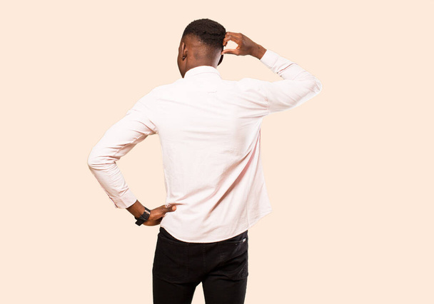young african american black man feeling clueless and confused, thinking a solution, with hand on hip and other on head, rear view against beige wall - Photo, image