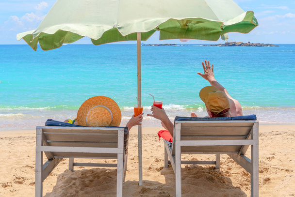 Ein Mann und eine Frau liegen mit bunten Cocktails in der Hand in Hängematten. winkt ein Mann mit der Hand zum Meer. Hallo Sommer. Hände mit bunten Cocktails am Strand - Foto, Bild