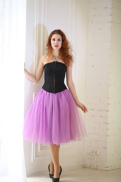 beautiful red-haired girl in a purple tulle skirt and black corset is standing near the floor window - Photo, Image