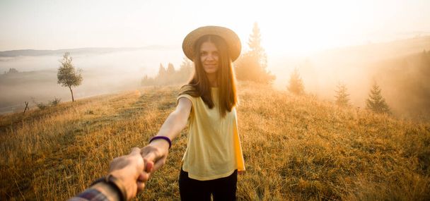 Ακολούθησέ με. Ζευγάρι ερωτευμένο κρατιέται χέρι χέρι. Γυναίκα σε κίτρινο πουκάμισο και ψάθινο καπέλο κρατώντας τον άνθρωπο με το χέρι πηγαίνει στο δάσος του φθινοπώρου με βουνά και συννεφιασμένο ουρανό το πρωί. - Φωτογραφία, εικόνα