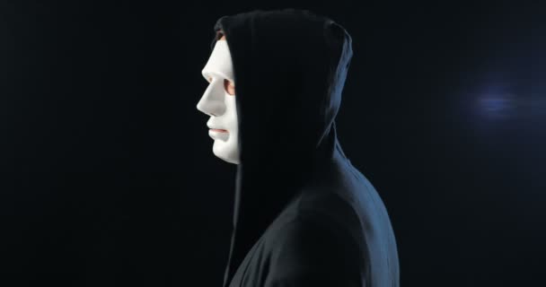 Anonieme man met een wit masker op zijn gezicht en een zwarte kap draait zich om en kijkt naar de camera. Zwarte donkere achtergrond. Begrip lid van een geheime organisatie - Video