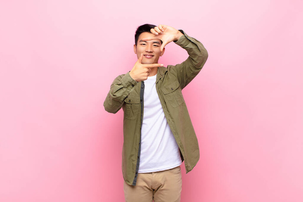 young chinese man feeling happy, friendly and positive, smiling and making a portrait or photo frame with hands against flat color wall - 写真・画像