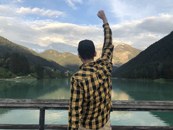 takaisin näkymä mies matkustava kohotettu käsi seisoo rannalla järven vuorten peitossa metsän päivällä
  - Valokuva, kuva
