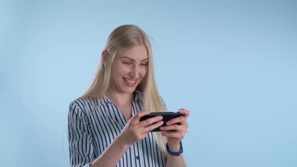 Pretty blonde girl playing games on smartphone on blue background - Záběry, video