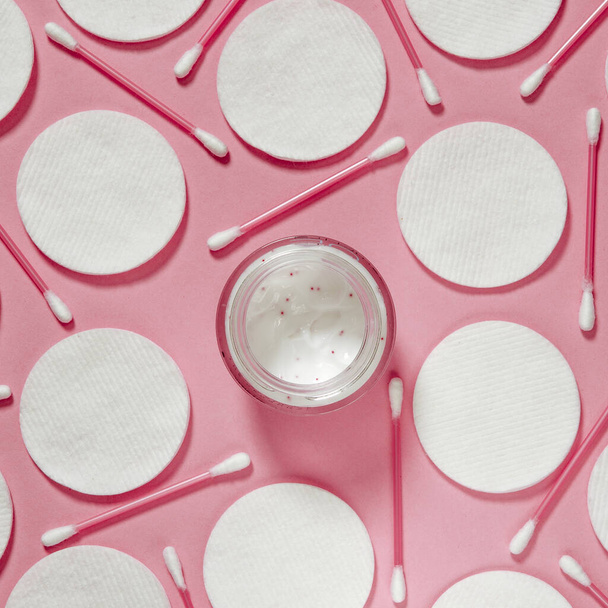 Hygiëneproducten: witte ronde wattenschijfjes en plastic sticks voor het reinigen van de oren liggen op een roze achtergrond naast gezichtscrème - Foto, afbeelding