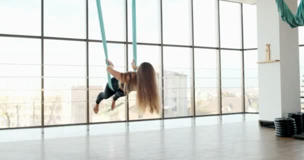 Beautiful young woman doing fly yoga exercise in the room with big windows. Attractive female working with her body in hammock. Relaxation concept - Footage, Video