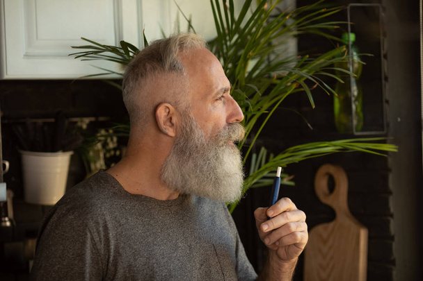 Bearded senior man smoking a cigarette at home. - Zdjęcie, obraz