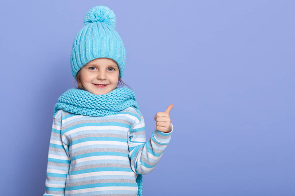 Positive attractive little girl with pleasant appearance dresses cap, scarf and shirt, points aside with her thumb, has cheerful facial expression, shows something at copy space. Advertisement concept - Fotó, kép