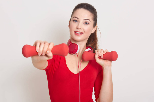 Pretty sporty woman using red dumbbells for her workout, listens to music while exercising, wears stylish sportwear, poses isolated over white background in studio, dark haired female looks at camera. - Fotografie, Obrázek