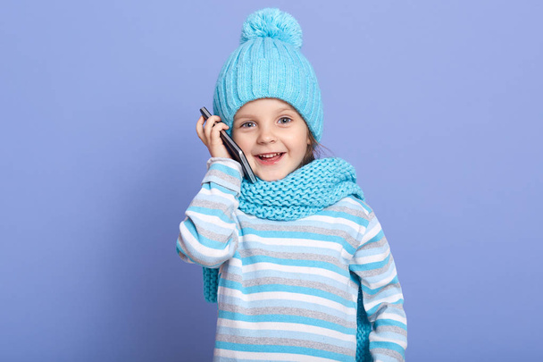 Happy little girl talking to her mother via modern cell phone against blue studio background, child having joyful facial expressing, being in good mood, dresses striped shirt, scarf and winter cap. - Photo, Image