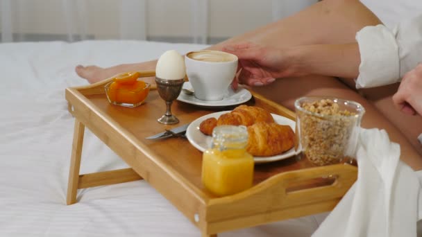 Close-up tiro de jovem bebendo café no quarto de hotel acolhedor pela manhã. Bandeja de mesa na cama branca com ovo, xícara, croissants. compota, mel, muesli e sumo de cenoura. Pequeno-almoço saudável. Tiros em 4K
 - Filmagem, Vídeo