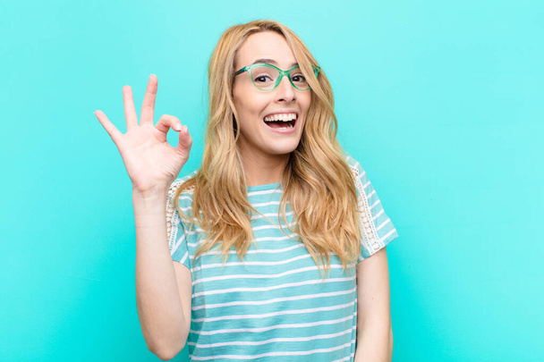 young pretty blonde woman feeling successful and satisfied, smiling with mouth wide open, making okay sign with hand against flat color wall - Φωτογραφία, εικόνα
