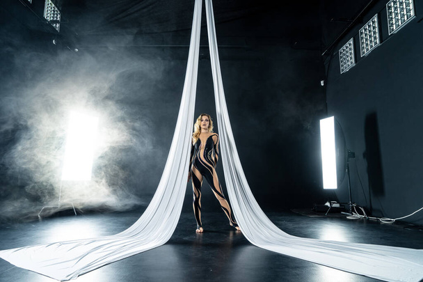 Beautiful flexible woman gymnast with white aerial silks isolated on black background with smoke - Фото, зображення