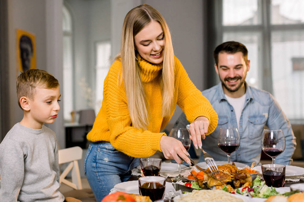 Portrait of happy couple parents and their little son sitting at festive table and going to eat roasted turkey. Happy family having delicious dinner together at home while mother cutting turkey - 写真・画像