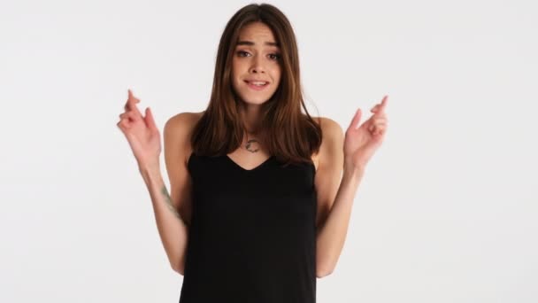 Attractive brown haired girl with crossed fingers hopefully praying on camera over white background. Please expression - Footage, Video