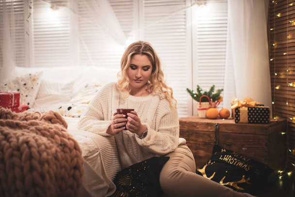 blonde girl in a sweater with a Cup of tea sitting on the bed - Fotó, kép