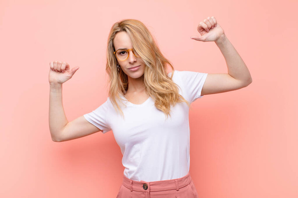 young pretty blonde woman feeling proud, arrogant and confident, looking satisfied and successful, pointing to self against flat color wall - Photo, Image