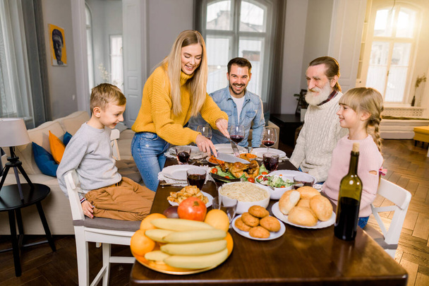 Family of five people, grandfather, parents and children sitting at the table and going to eat roasted turkey, while happy mother is cutting turkey. Thanksgiving family dinner concept - 写真・画像