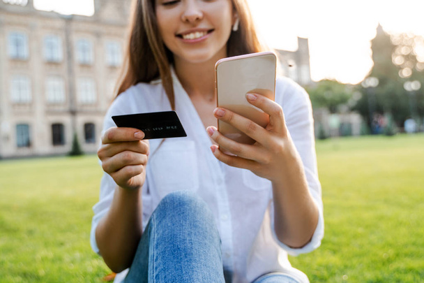 Using credit card and making purchases online via internet facilities, cash back offer by shopping in online platform. Happy user woman buy or booking something and smiling. Sitting on green grass - Photo, Image