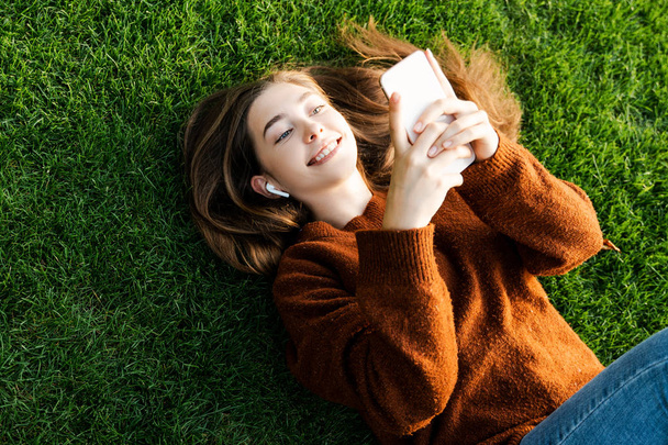 Lächelnd fröhliches Teenager-Mädchen, das auf ihr Smartphone schaut und Freunde anruft, um online zu kommunizieren, mit moderner digitaler Online- und Internettechnologie, entspannt auf grünem Gras liegend - Foto, Bild