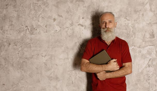 Bearded Senior man stands on grey wall and holding a book. - Фото, изображение