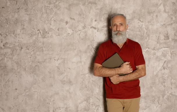 Homme âgé barbu se tient sur le mur gris et tenant un livre
. - Photo, image