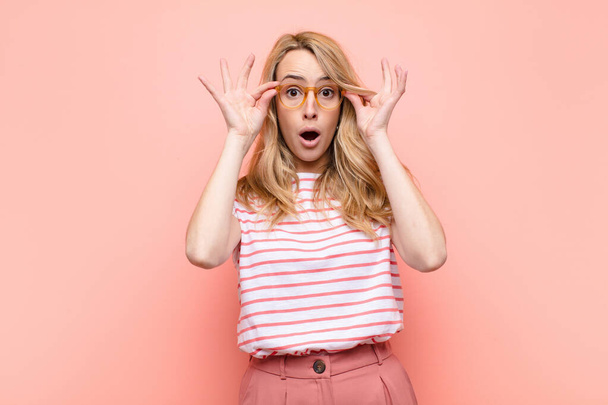 young pretty blonde woman feeling shocked, amazed and surprised, holding glasses with astonished, disbelieving look against flat color wall - Foto, Imagen