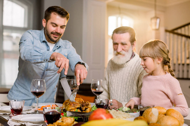 Padre sonriente cortando rebanadas de pavo para la cena familiar. Familia con niños comiendo cena de Acción de Gracias. Pavo asado en mesa de comedor
. - Foto, imagen