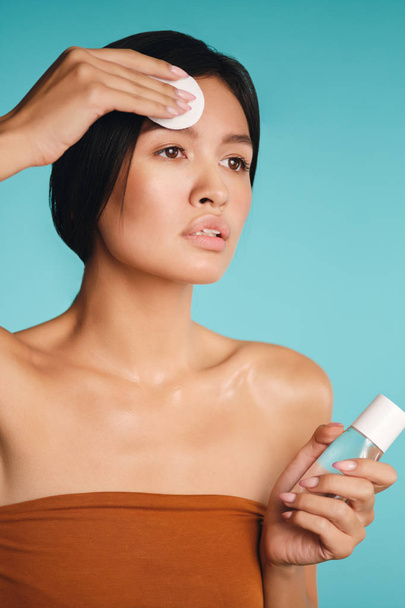 Aantrekkelijk aziatisch brunette meisje aandachtig op zoek weg schoonmaken gezicht met spons over kleurrijke achtergrond - Foto, afbeelding