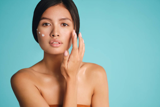 Красивая азиатская брюнетка с точками крема на щеке чувственно глядя в камеру на красочном фоне
 - Фото, изображение