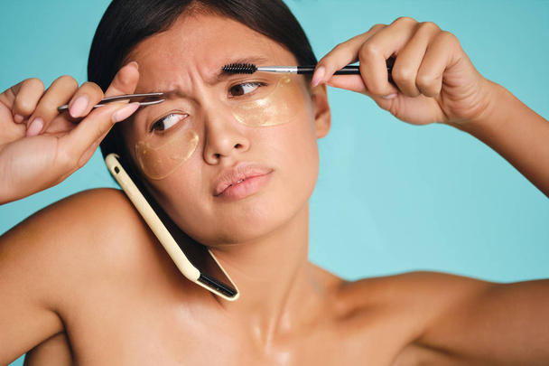 Busy Asian girl with patches under eyes putting cellphone between head and shoulder while using tweezers and brow brush over colorful background - Photo, Image