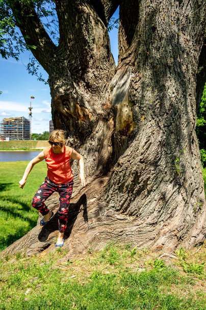 Mujer madura en rojo descansando cerca de un enorme árbol junto al río
 - Foto, Imagen