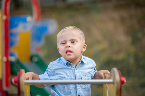 Η καθημερινή ζωή ενός παιδιού με αναπηρίες. Ένα αγόρι με σύνδρομο Ντάουν παίζει στην παιδική χαρά. Χρωμοσωμική και γενετική διαταραχή στο μωρό. - Φωτογραφία, εικόνα
