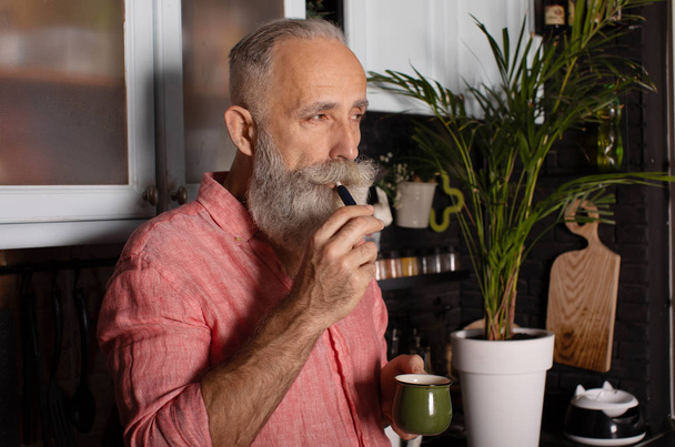 Front view of bearded senior man with coffee cup on his kitchen - Fotó, kép