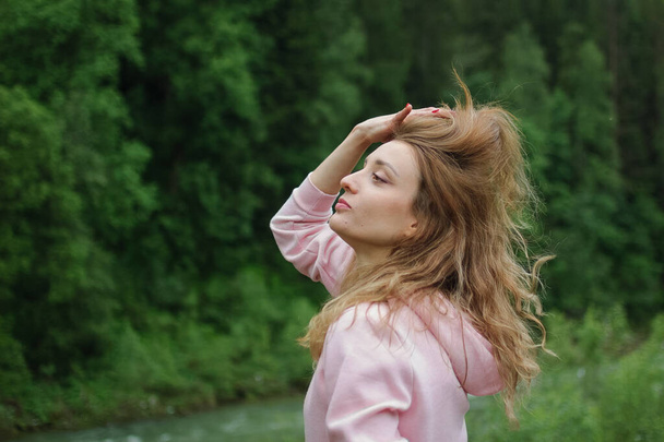 Вид сбоку удивительной девушки в розовой повседневной одежде с длинными волосами и очаровательной улыбкой на холме на зеленом фоне леса весной или ранней осенью в горах
. - Фото, изображение