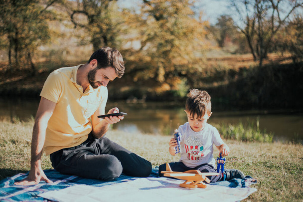 Een drie jaar oude jongen speelt op een picknick in de buurt van zijn oom die kijkt naar de telefoon - Foto, afbeelding