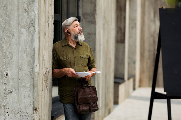 A gray-haired, bearded Manager talks wirelessly, and holds business documents in his hands - Fotó, kép
