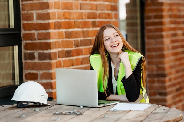 A red haired architect girl sits in an office in a yellow vest and looks away with a smile - Foto, Imagem