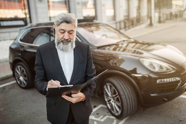 A grizzled adult used car salesman waits for a buyer next to a representative car - 写真・画像