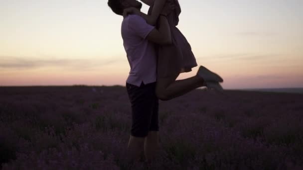 Sylwetka Młody mężczyzna podnosi swoją dziewczynę w ramionach stojąc na kwitnącym polu lawendy o zachodzie słońca - Materiał filmowy, wideo