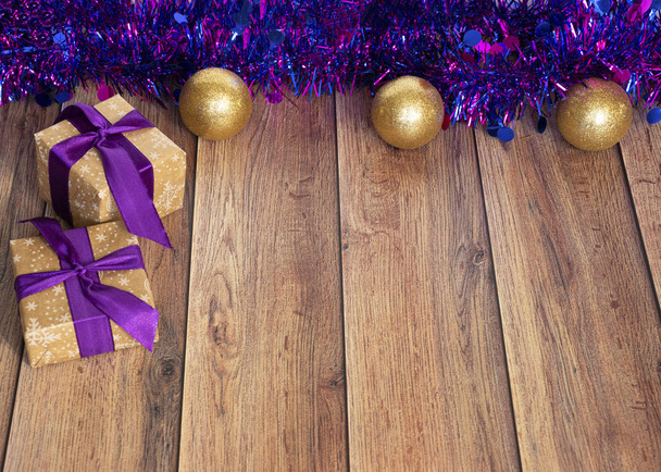 Τα δώρα είναι συσκευασμένα σε χαρτί Kraft και δεμένα με σατέν κορδέλα με χριστουγεννιάτικα παιχνίδια και μωβ tinsel σε ξύλινο φόντο. Έκπληξη για τις διακοπές. Διακόσμηση για το χριστουγεννιάτικο δέντρο - Φωτογραφία, εικόνα