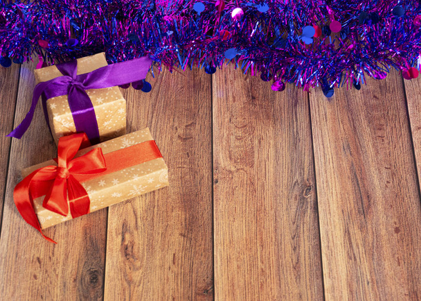 Подарки упакованы в крафтовую бумагу и завязаны атласной лентой с новогодними игрушками и мишурой на деревянном фоне. Сюрприз для праздника. Декор для елки
 - Фото, изображение