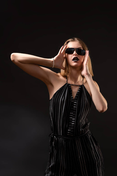 Fiatal, gyönyörű nő fekete overálban és napszemüvegben, fekete háttér felett pózolva, felülnézetből. Stúdió lövés barna nő ellen elszigetelt háttér - Fotó, kép