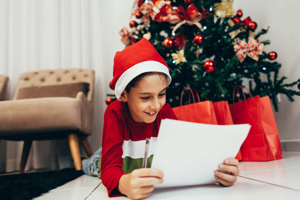 Boy lying on the floor under the Christmas tree writing wish list for Santa. Waiting for Christmas. Celebration. New Year. - Φωτογραφία, εικόνα