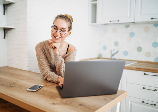 Bela sorridente jovem mulher clara cabelo longo menina em óculos vestindo em camisola de malha aconchegante usando laptop na cozinha brilhante
 - Foto, Imagem