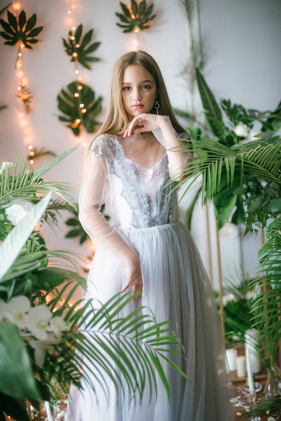 Attractive young girl with blond hair and evening make-up in a romantic dress among tropical greenery and white orchids, roses and lilies. Beauty and fashion - Фото, изображение