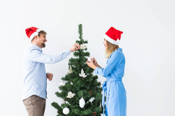 Holidays and festive concept - Young family couple in santa hats decorating the christmas tree - Photo, Image